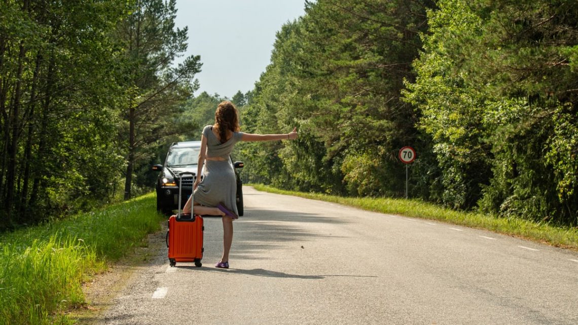 A woman hitchhiking with an orange suitcase on a rural road in Estonia during summer.