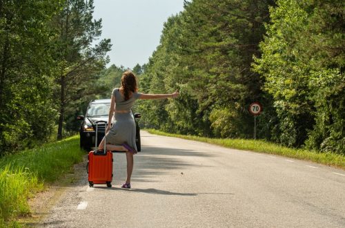 A woman hitchhiking with an orange suitcase on a rural road in Estonia during summer.