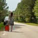 A woman hitchhiking with an orange suitcase on a rural road in Estonia during summer.